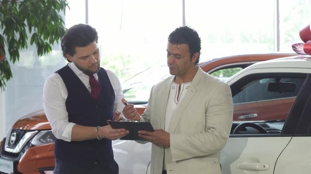 Handsome mature man buying a new automobile at the dealership, signing papers with the salesman. Professional car dealer selling a new automobile to a male customer. Retail, sales.