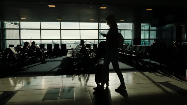 A woman with a bag on wheels and a phone in her hand is walking along the airport terminal. Silhouet against the background of large windows