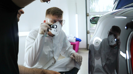 Professional master after polishing and painting cars, checks the flashlight scratches, a respirator, white rubber gloves, white coat, goggles, auto service.	