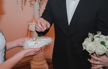 Couple in love is getting married. Groom in black suit is taking golden ring and wearing it on bride's finger. Bride is holding cute white wedding bouquet in hands.