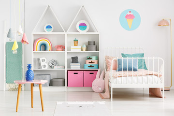 Front view of a kid's room with a table, shelves with boxes and rainbow, single bed and ice cream poster on the wall - obrazy, fototapety, plakaty