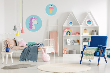 Colorful kid's bedroom interior with a unicorn and ice cream poster, bed with sheets, rabbit pillow, shelves and blue armchair with a blanket