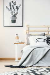 Cactus poster on white wall above bed with grey sheets in bright bedroom interior. Real photo