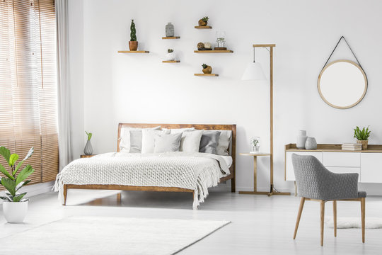 Front view of a bright natural bedroom interior with wooden bed with cover and pillows, nightstand and lamp, gray armchair and round mirror on white wall. Real photo.