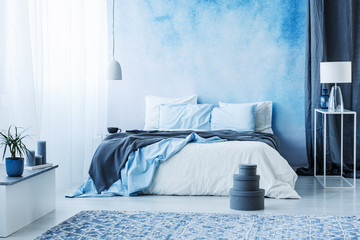 Grey boxes next to bed with blue bedding in bedroom interior with white lamp against a curtain - obrazy, fototapety, plakaty