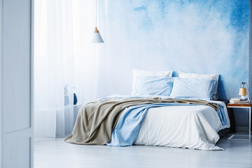 Yellow and blue bedding on white bed in minimal bedroom interior with lamp on wooden table