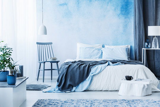 Blue chair next to bed with grey blanket in cozy bedroom interior with plants on white cupboard