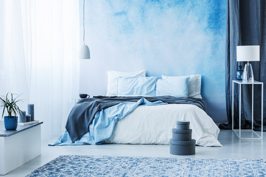 Grey boxes next to bed with blue bedding in bedroom interior with white lamp against a curtain