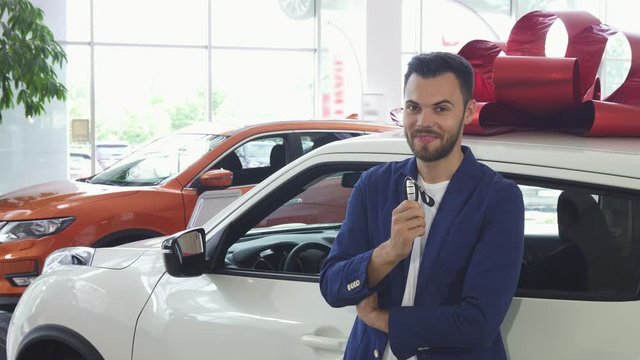 Shot of a handsome young man smiling joyfully, leaning on his new automobile, showing car keys to the camera. Ownership, driving, travelling concept. Man buying auto at the dealership.