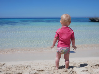 Little baby in pink swimming suit looking at the aquamarine blue see from a sand beach of Favignana island (Sicily, Italy).