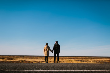 A young couple of travelers stands together on empty road on backgrouns of blue sky. Rear view. Iceland traveling