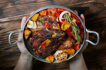 Hands holding fried ribs in a pan with rosemary, potatoes rustic, onion, sauce . Ribs are strung on a meat fork. Dark background. Place for text, copyspace