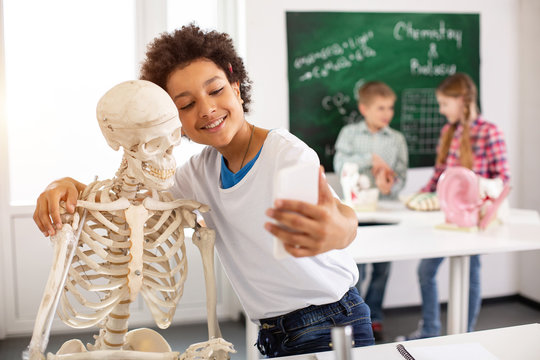 Positive mood. Nice cheerful boy hugging a skeleton while taking selfies with him