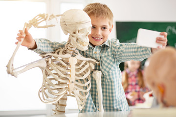 So cool. Cheerful nice boy holding a skeletons hand while taking a photo with it