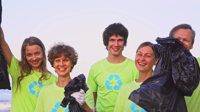 Five young volunteers in green t-shirts with image recycle, collect garbage on an oceanic beach, then pose on the camera with bags of collected garbage. Volunteering and recycling concept