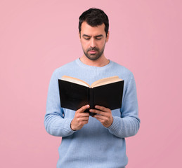 Man with blue sweater holding and reading a book on pink background