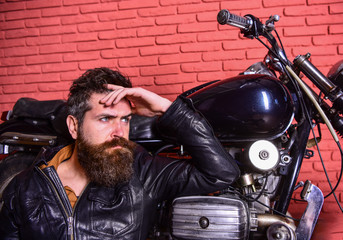 Fototapeta na wymiar Bikers lifestyle concept. Man with beard, biker in leather jacket near motor bike in garage, brick wall background. Hipster, brutal biker on pensive face in leather jacket sits, leans on motorcycle.