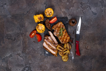 sliced pork steak and grilled vegetables on slate board on stone table.