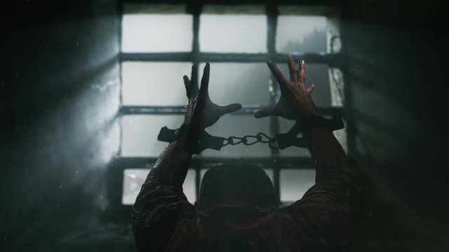 Prisoner with chained hands pray to the Lord. View trough prison window. Dolly shot.