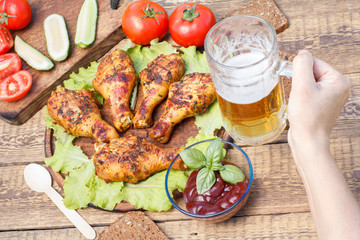 Grilled chicken legs and lettuce leaves on wooden chopping board, tomato sauce in glass bowl and female hand holding glass mug of beer