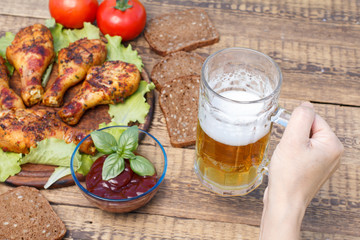 Female hand is holding glass mug of beer with grilled chicken legs and tomato sauce on the background