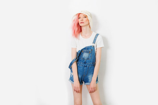 fashion sexy hipster summer girl with pink hair in jeans overalls and hat posing near white urban wall along