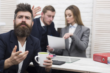 Man with beard on hopeful face holds mug, bosses, coworkers, colleagues on background. Business negotiations concept. Man hope for success, waiting while partners discuss contract.