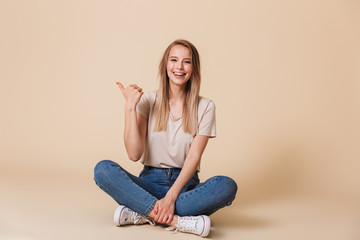Photo of beautiful woman pointing finger aside at copyspace while sitting on floor with legs crossed, isolated over beige background