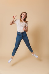 Full length portrait of excited blond woman 20s wearing casual clothing jumping and showing peace sign with two hands, isolated over beige background