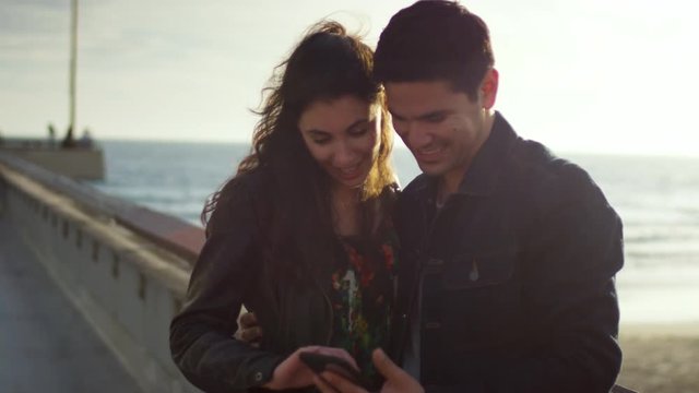 Happy Couple Swipe Through Their Vacation Photos On Smartphone, At Pier, Beautiful Sunset (Slow Motion)