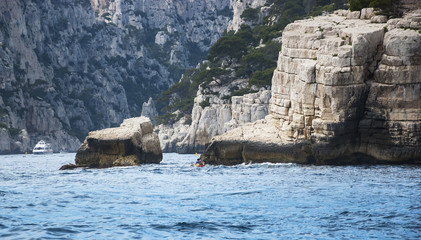 Three men row in the canoe between the rocks in the famous Calanques national park of Cassis (near Marseilles in Provence, France) and tourist ships at backgrounds.