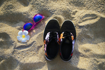 Naklejka premium Sneakers with sunglasses and flower on the beach sand 