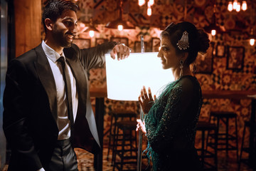 Smiling Indian wedding couple in European evening dresses stand before a bright lamp in dark room