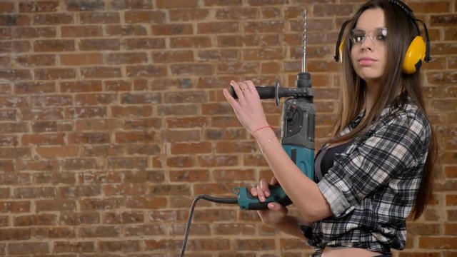 Beautiful attractive young women in glasses and headphones holding perforator and looking into camera, female builder, brick background