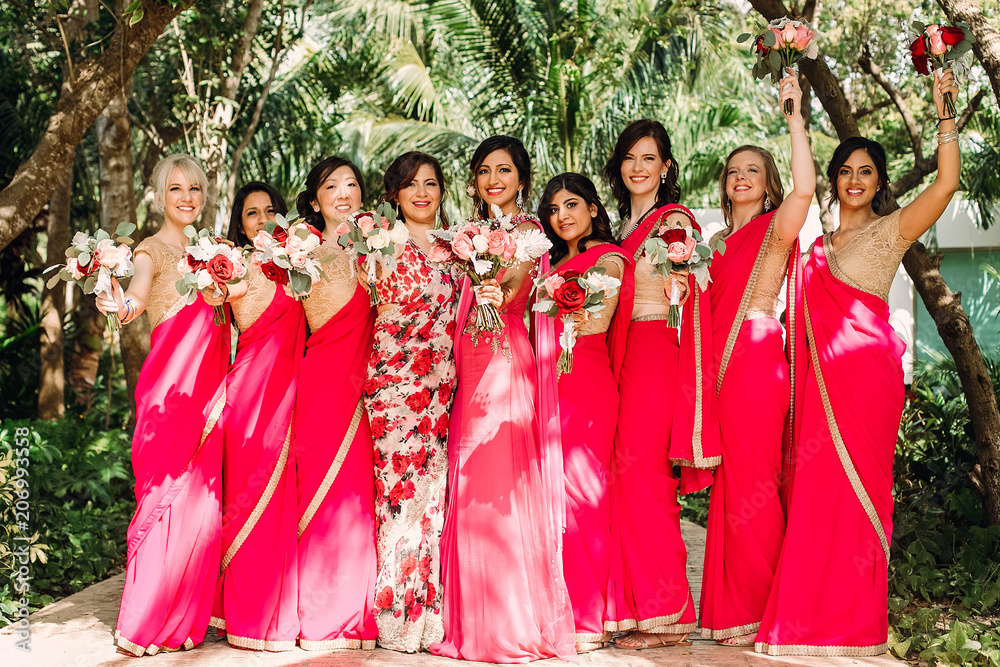 Wall mural Pretty Indian bridesmaids in red sari stand around beautiful bride in pink sari posing outside