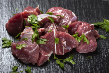 Pieces of raw beef meat on a dark slate with green herbs