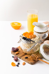 Healthy breakfast in glass mason jar with oat flakes, granola, milk, dry fruits and nuts, dates, honey, orange juice, eggs for morning fitness on wooden board platter on white background isolated