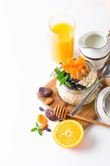 Healthy breakfast in glass mason jar with oat flakes, granola, milk, dry fruits and nuts, dates, honey, orange juice, eggs for morning fitness on wooden board platter on white background isolated