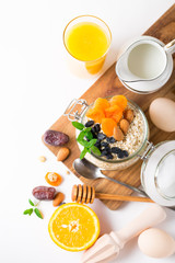 Healthy breakfast in glass mason jar with oat flakes, granola, milk, dry fruits and nuts, dates, honey, orange juice, eggs for morning fitness on wooden board platter on white background isolated