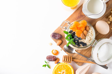 Healthy breakfast in glass mason jar with oat flakes, granola, milk, dry fruits and nuts, dates, honey, orange juice, eggs for morning fitness on wooden board platter on white background isolated