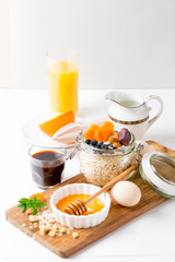 Healthy breakfast in glass mason jar with oat flakes, granola, milk, dry fruits and nuts, dates, honey, orange juice, eggs for morning fitness on wooden board platter on white background isolated