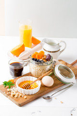 Healthy breakfast in glass mason jar with oat flakes, granola, milk, dry fruits and nuts, dates, honey, orange juice, eggs for morning fitness on wooden board platter on white background isolated