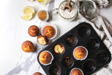 Fresh baked homemade lemon cakes muffins standing in teflon baking dish with eucalyptus branch, linen cloth and ingredients in glass jars above over white marble texture background. Flat lay, space