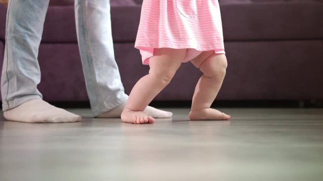 Little baby feet walking on floor with parent support. Close up of mother learning child walking. Kid feet steps at home. Little child learn to walking. Baby steps with mom. Toddler doing first step
