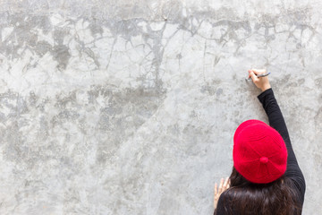 Beautiful woman is drawing or writing some text, picture or commercial by using a pen. Gorgeous woman writes on beautiful cement wall. Pretty girl wears red hat, long sleeves shirt. wide copy space