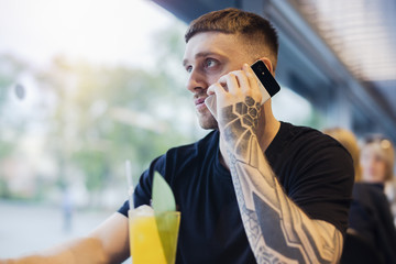 Attractive young man with tattoo is holding mobile phone and drinking lemonade.