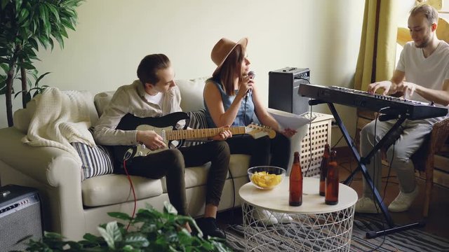Musical band is rehearsing performance singing and playing musical instruments in home studio looking at music sheet. Bottles and snacks on table are visible.