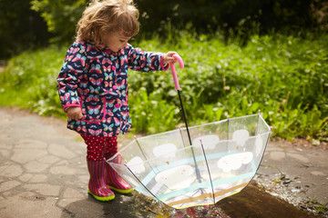 Charming little girl with umbrella has fun standing in gumboots in the pool after rain