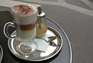 Close up of a clear tall glass coffee cup with layered latte topped with cinnamon on silver plate, outdoor cafe, Cappuccino