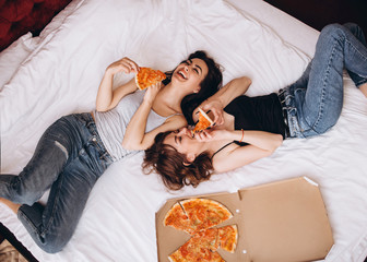 The stylish sisters eating slices of pizza and lying on the bed